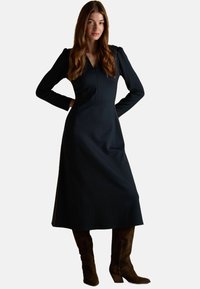 Must mustaga mustriga kleit V-kaelusega, struktureeritud õlgadega ja voolava seelikuosaga; kombineeritud pruunide sametist pahkluu saapadega.