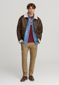 Brązowa skórzana kurtka z wełnianym kołnierzem, jasnoniebieska koszula dżinsowa, bordowa koszulka, khaki spodnie cargo i brązowe buty na sznurówki.