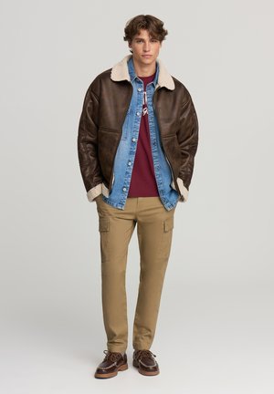 Giacca di pelle marrone con collo in shearling, camicia di jeans azzurro chiaro, T-shirt bordeaux, pantaloni cargo color cachi e scarpe marroni stringate.