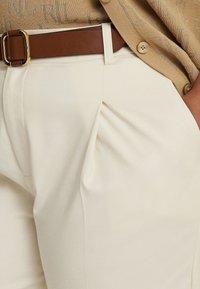 Lähikuva vaaleista pleidetud püksidest pruunist nahavööst ja beežist nööpidest topist, mis on sisse pandud.