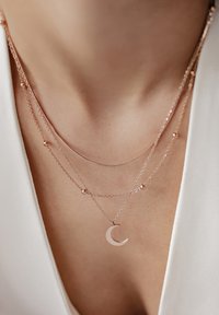 Colliers en couches en or rose avec un pendentif en forme de croissant de lune, des accents perlés et des chaînes délicates sur un fond neutre.