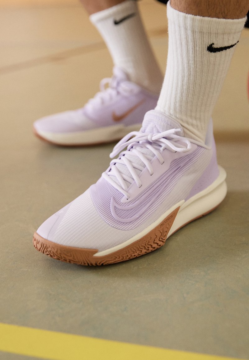 Nærbillede af fødder iført hvide Nike-sokker og lyse lilla Nike-sneakers på et gymgulv med gule bane-linjer.
