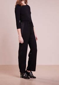 Schwarzer Overall mit langen Ärmeln, elastischem Bund und Kordelzug. Verfügt über eine lockere Passform und leicht ausgestellte Beine. Kombiniert mit schwarzen Absatzschuhen.