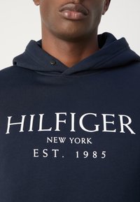 Gros plan d'une personne portant un sweat à capuche bleu marine avec le texte blanc « HILFIGER NEW YORK EST. 1985 » à l'avant.