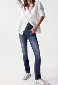 Jeans de mezclilla azul oscuro con detalles desgastados, talle alto y cierre de botones. Combinados con una camisa de lino blanca de botones y zapatillas blancas.