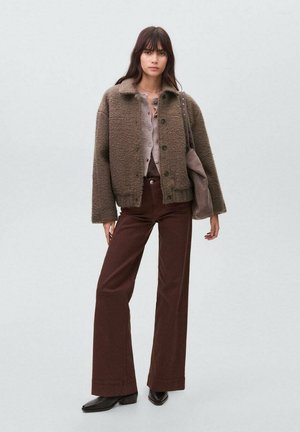 Veste en laine texturée de couleur marron avec un col, boutons noirs, associée à un pantalon évasé marron et des bottes à talons noires. Palette de couleurs terreuses.