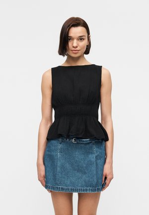 Kobieta z krótkimi ciemnymi włosami, ubrana w czarny, bezrękawnikowy top z baskinką i niebieską dżinsową mini spódniczkę z paskiem, stojąca na jednolitym tle.