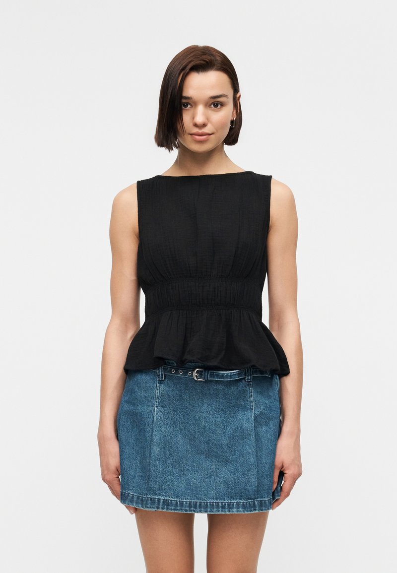 Γυναίκα με κοντά σκούρα μαλλιά, φορώντας μαύρο αμάνικο τοπ με πέπλουμ και μπλε μίνι φούστα από τζιν με ζώνη, στέκεται μπροστά σε απλό φόντο.