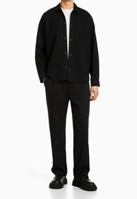 Svart skjorta med knappar och ficka, strukturerad tyg, ihopparad med svarta byxor och kraftiga svarta skor, vilket skapar en monokrom outfit.