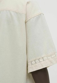 Camisa de lino color beige claro con corte holgado, mangas cortas y detalle de encaje decorativo en el dobladillo de las mangas. Textura suave, diseño minimalista.