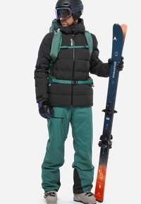 Schwarze, voluminöse Jacke mit grünen Akzenten, türkisfarbene Skihose und ein Trinkrucksack. Hält Skier, trägt einen schwarzen Helm und spiegelnde Goggles.