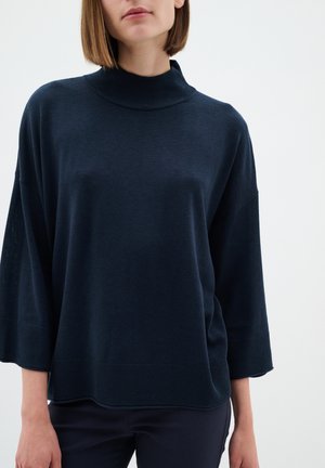 Navy blue neulepusero, jossa on korkea kaulus, alemmat olkaset ja leveät hihat. Varustettu ribbireunoilla hihoissa ja helmassa. Pehmeä rakenne.