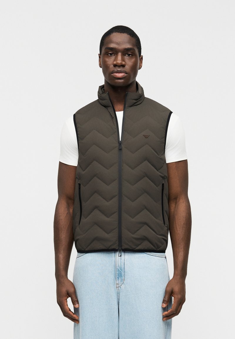 Άνδρας που φοράει αμάνικο γιλέκο σ ε σκούρο ελιά πράσινο χρώμα με καπιτονέ ύφασμα πάνω από άσπρο κοντομάνικο πουκάμισο και ανοιχτόχρωμο τζιν, που στέκεται μπροστά σε απλό λευκό φόντο.