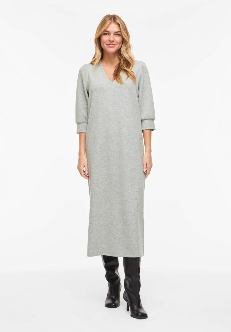 Femme aux longs cheveux blonds portant une robe midi gris clair à col en V et des bottes noires à talons montant jusqu'aux genoux, debout devant un fond blanc.