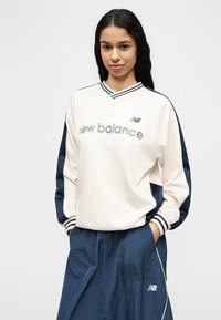 Νεαρή γυναίκα με μακριά σκούρα μαλλιά, που φοράει μπεζ και ναυτικό μπλε φούτερ New Balance και ναυτικό μπλε αθλητικό παντελόνι, με τα χέρια στις τσέπες.