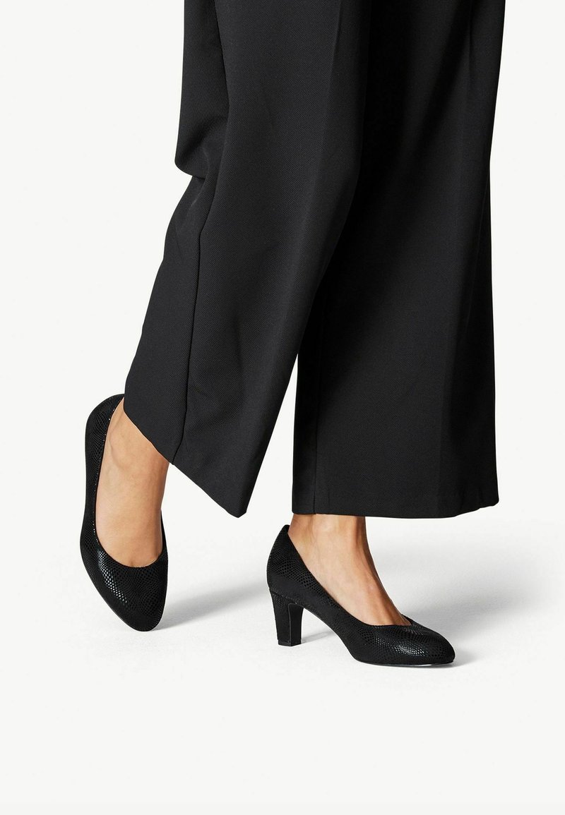 Fötter som bär svarta låga stylter med struktur, parades med svarta vida byxor på vit bakgrund.