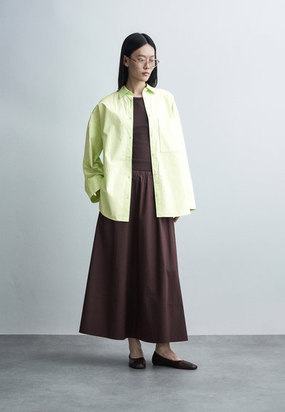 Femme aux longs cheveux noirs portant des lunettes transparentes, une chemise ample jaune clair boutonnée sur une robe marron foncé, et des chaussures plates foncées, debout à l'intérieur.