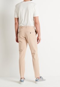 Beige snugga byxor med en slät yta, två bakfickor med svarta knappar och en elegant siluett, matchade med grå sneakers.