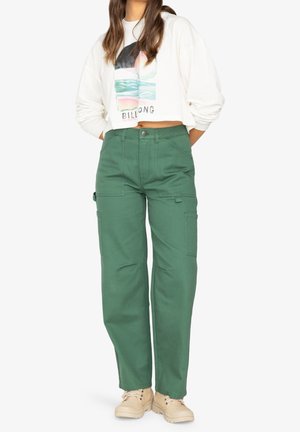 Kobieta w zielonych spodniach cargo, beżowych butach i białej krótce przyciętej bluzie z wielokolorowym logo Billabong na białym tle.
