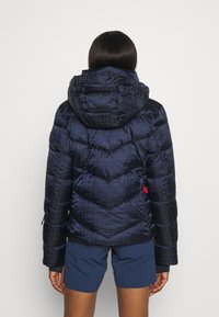 Tmavě modrá prošívaná bunda s kapucí, zdobená vzorem šipek a vystouplým vzorem „FIRE“. Obsahuje červené detaily a kapsy na bocích.