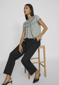 Jasnozielona bluzka z falbanami, w połączeniu z czarnymi spodniami o szerokich nogawkach i czarnymi szpilkami, siedząca na drewnianym stołku.