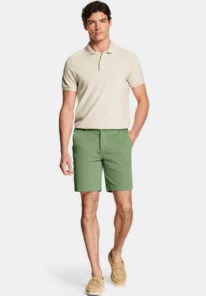 Uomo con polo beige a maniche corte, pantaloncini verdi e scarpe da barca color cuoio, in piedi con una mano in tasca contro sfondo bianco.