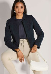 Vrouw in een getextureerd zwart blazer, marineblauwe blouse, beige broek, met een witte handtas, zittend tegen een effen lichte achtergrond.