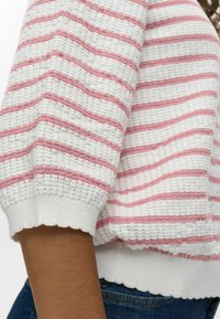 Csíkos pulóver fehér és rózsaszín csíkokkal, textúrált anyagból, fodros mandzsettákkal és laza szabásban. Közeli fotó a ujjak részleteiről.