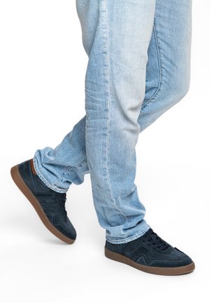 Osoba v světlých modrých džínách a černých teniskách s hnědými podrážkami stojí s jednou nohou přes druhou.