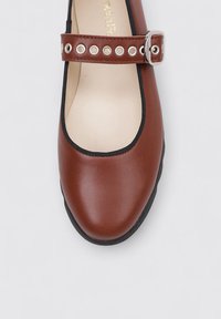 Zapato plano de cuero marrón con punta redonda, ribete elástico negro y una correa con ojales de metal y detalle de hebilla plateada.