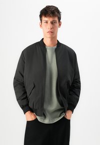 Junger Mann mit kurzen braunen Haaren, der eine schwarze Bomberjacke über einem hellgrünen Hemd trägt, die Hände in den Taschen, vor einem weißen Hintergrund.