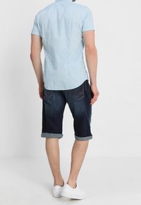 Camisa de manga corta azul claro con un patrón sutil, combinada con pantalones cortos de mezclilla oscura doblados en el dobladillo y zapatillas blancas.