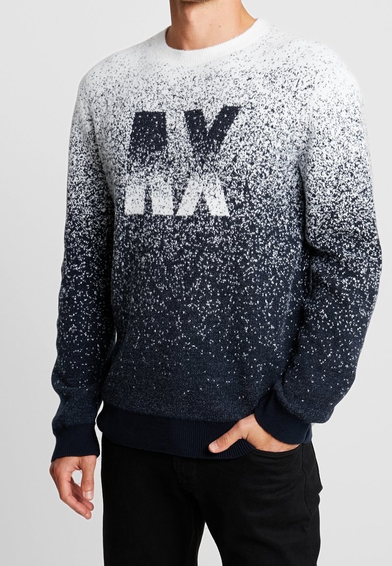 Man som bär en marinblå och vit prickig tröja med stora "AX" bokstäver, ihop med svarta byxor, med handen i fickan, mot en enfärgad bakgrund.