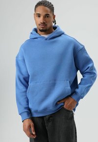 Blauwe hoodie gemaakt van zachte stof met een kangoeroezak, voorzien van geribbelde boorden en zoom. Model draagt deze met een donkerblauwe spijkerbroek.