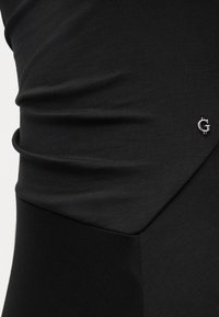 Κοντινό πλάνο μαύρου υφάσματος με ρυτιδώσεις και μικρή ασημένια διακόσμηση με το γράμμα "G" σε εφαρμοστό ρούχο.