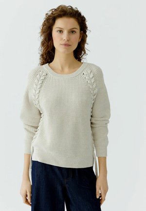 Femme aux cheveux bouclés portant un pull en maille crème avec des détails tressés et un jean bleu foncé, debout devant un fond clair uni.