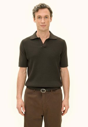 Man met kort krullend haar die een donkerbruin, gestructureerd poloshirt met korte mouwen en een bruine broek met zwarte riem draagt, staand tegen een lichte achtergrond.