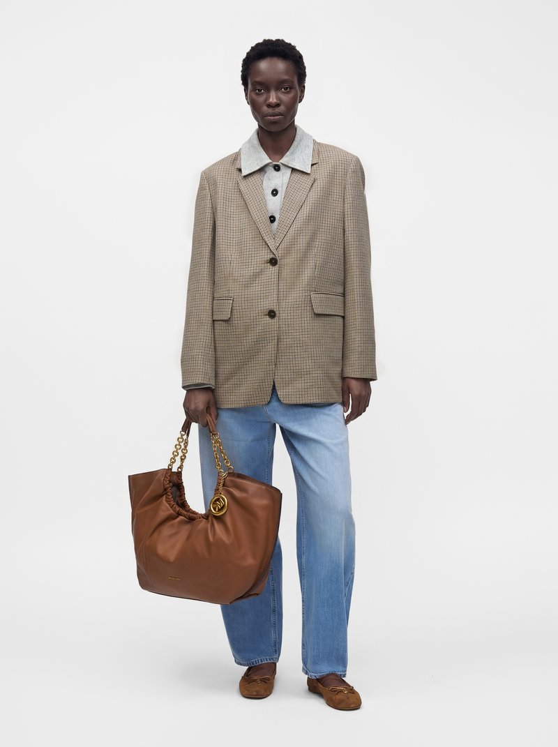 Femme portant un blazer beige à carreaux, une chemise grise boutonnée, un jean bleu clair, des mocassins marron, tenant un grand sac à main en cuir marron avec des anses chaînes dorées.