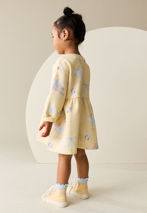 Jeune fille avec les cheveux en chignon portant une robe jaune avec des imprimés de fée et de papillon et des baskets montantes jaunes assorties avec des chaussettes bleues à volants.
