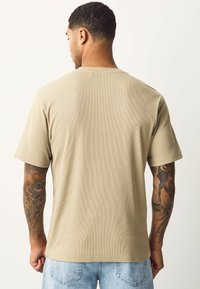 Camiseta beige de manga corta con tejido texturizado, corte relajado y cuello redondo. El modelo lleva pantalones cortos de mezclilla de lavado claro y tiene los brazos tatuados.
