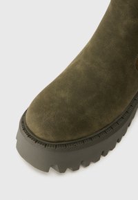 Primo piano di un stivale in suede verde oliva con una spessa suola in gomma nera e un motivo di battistrada robusto su sfondo bianco.