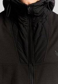 Zwarte jas met hoge kraag, voorzien van een gewatteerde textuur, dubbele ritsluiting en een klein logo detail aan de zijkant.