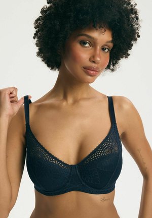 Žena s kučeravými vlasmi si upravuje ramienko námornícky modrej čipkovanej podprsenky, ukazuje horný trup a tetovania na rukách.