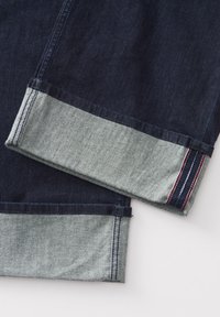 Tessuto denim scuro con un polsino in denim chiaro a contrasto. Il polsino presenta dettagli di cucitura rossi e bianchi. Texture liscia, forma dritta.