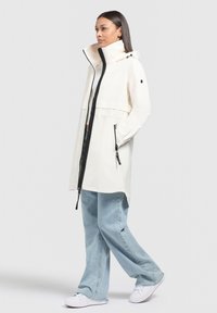 Giacca impermeabile bianca con colletto alto, zip nera e accenti. Abbinata a jeans in denim azzurro chiaro a gamba larga e sneakers bianche.