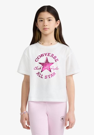 Jong persoon met lang donker haar die een wit Converse All Star T-shirt met een roze ster en lichtroze broek draagt tegen een witte achtergrond.