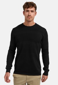 Maglione nero lavorato a maglia con strisce strutturate, scollatura rotonda e maniche lunghe, abbinato a pantaloni di colore chiaro. Nessun marchio visibile.