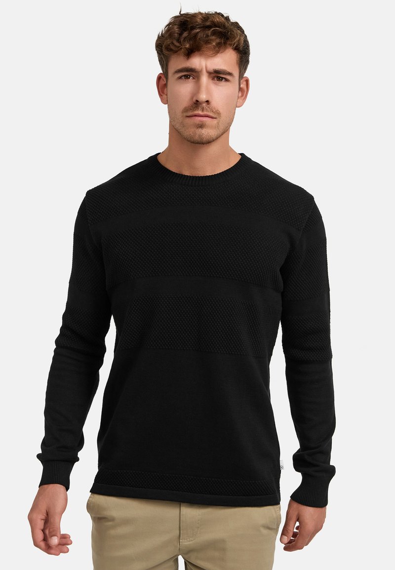 Schwarzer Strickpullover mit strukturierten Streifen, rundem Ausschnitt und langen Ärmeln, kombiniert mit hellen Hosen. Keine sichtbare Markenkennzeichnung.
