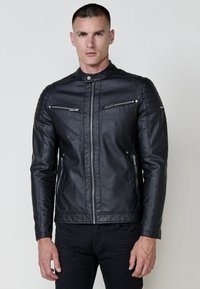 Homme portant une veste en cuir noir ajustée avec des poches zippées sur la poitrine et sur les côtés, se tenant devant un fond clair uni.