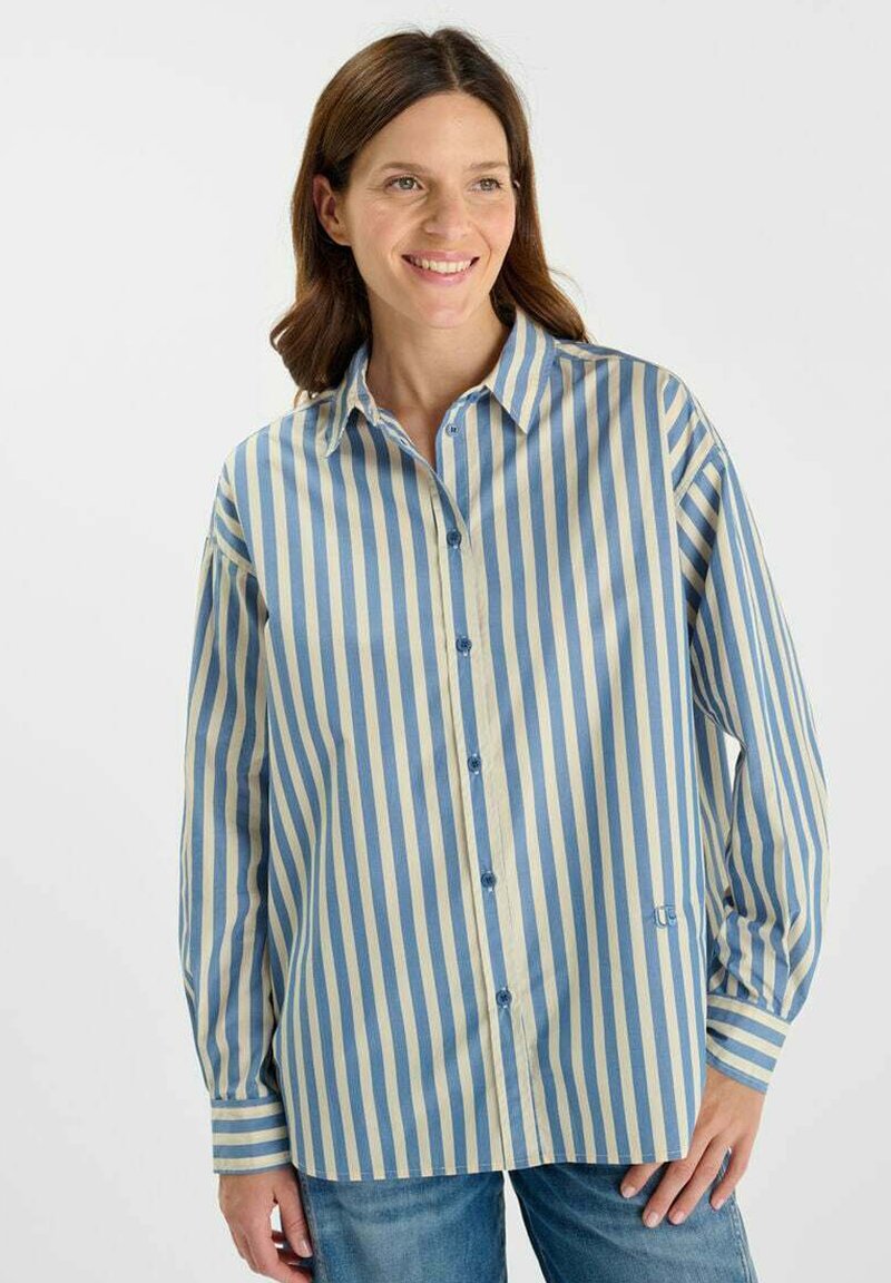 Donna sorridente con capelli castani di lunghezza fino alle spalle, che indossa una camicia a pulsante blu e beige a righe verticali e jeans blu.